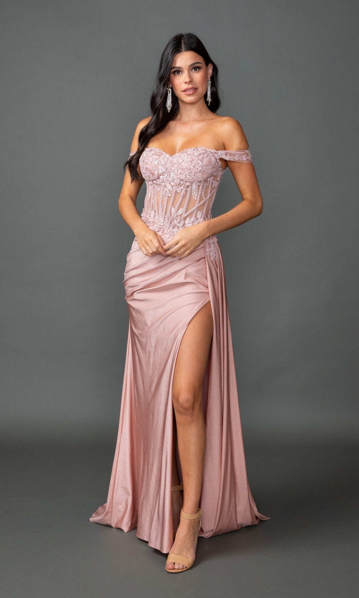 A woman stands against a gray background wearing the Dancing Queen 4535, a shimmery blush pink off-the-shoulder long prom dress with lace embroidery on the sweetheart neckline, a sheer corset bodice, and a high leg slit.